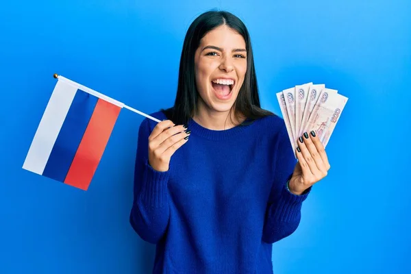 年轻的惊慌失措的女人举着俄罗斯国旗和卢布钞票 笑着大声笑着 因为有一个可笑的疯狂笑话 — 图库照片