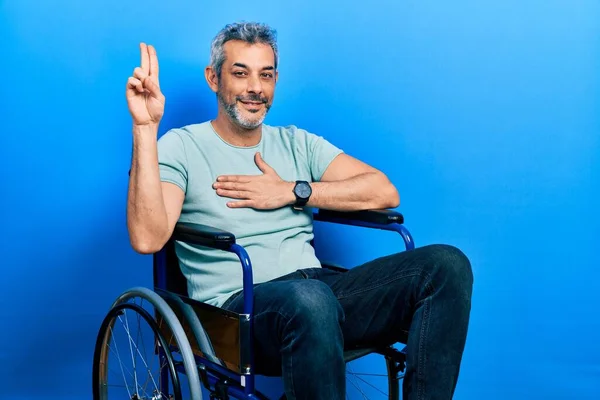 Handsome middle age man with grey hair sitting on wheelchair smiling swearing with hand on chest and fingers up, making a loyalty promise oath