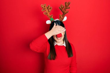 Geyik Noel şapkası takan, kırmızı burunlu, gülümseyen ve gülen genç İspanyol kadın şaşırmak için gözlerini kapatıyor. kör kavram. 