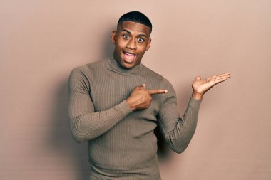 Balıkçı yaka kazak giyen genç siyah adam kameraya hayret ediyor ve gülümsüyor.. 