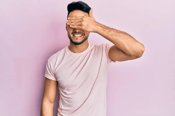 カジュアルな服を着た若いアラブ人男性が笑顔で 驚きのために目を覆う顔で手で笑っています 視覚障害の概念 — ストック写真