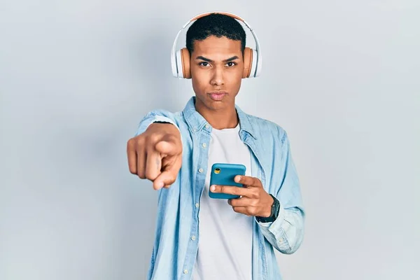 カメラとあなたを指差すヘッドフォンを身に着けているスマートフォンを使用して若いアフリカ系アメリカ人の男 真剣に見て自信を持ってジェスチャー — ストック写真