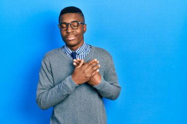 İş tarzı giyinmiş, gözlüklü, elleri göğsünde, gözleri kapalı gülümseyen ve yüzünde minnet ifadesi olan genç bir Afrikalı Amerikalı. sağlık kavramı. 