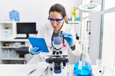 Laboratuvarda dokunmatik ped ve mikroskop kullanarak bilim adamı üniforması giyen İspanyol genç bir kadın.