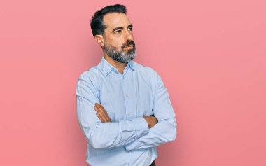 Orta yaşlı, sakallı, iş gömleği giyen, kolları çapraz ve kendinden emin bir şekilde yana bakan bir adam. 