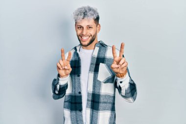 Modern boyalı saçlı, sıradan tişörtlü, gülümseyen, parmaklarını zafer işareti yaparken gösteren genç İspanyol bir adam. İki numara.. 