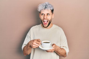 Modern boyalı saçları olan genç İspanyol bir adam bir fincan kahve içiyor kızgın ve kızgın çığlıklar atıyor sinirlenmiş ve öfkeli, öfkeyle bağırıyor.. 
