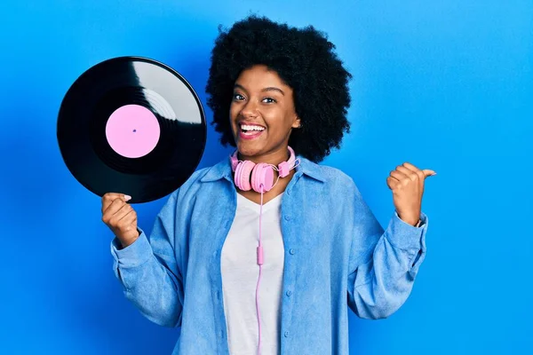 年轻的非洲裔美国女人戴着耳机 手里拿着乙烯基唱片 手指指向旁边 笑着张开了嘴 — 图库照片