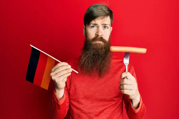 红头发的男人留着长长的胡子 手里拿着叉子 端着猪肉香肠和德国国旗 脸上带着严肃的表情 简单而自然地看着相机 — 图库照片