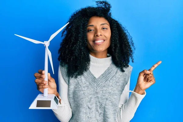 アフリカ系アメリカ人女性アフリカ系アメリカ人の髪の再生可能エネルギーのためのソーラー風車を保持笑顔幸せ側に手と指を指して — ストック写真