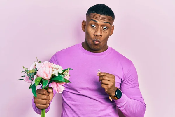 Young black man holding bouquet of flowers and wedding ring making fish face with mouth and squinting eyes, crazy and comical.