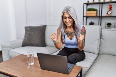 Orta yaşlı gri saçlı kadın evinde dizüstü bilgisayar kullanıyor. Kolları havada ve gözleri kapalı zaferi kutlarken gülümsüyor. Kazanan konsept. 