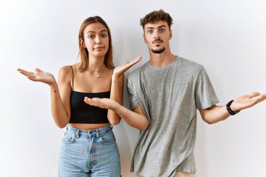 İzole edilmiş arka planda yan yana duran genç ve güzel bir çift. Kolları ve elleri havada, kafası karışmış bir ifade. şüphe kavramı. 