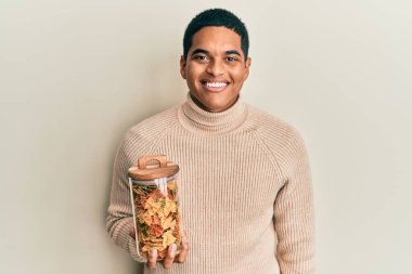 Genç, yakışıklı İspanyol adam elinde farfalle makarna kavanozuyla pozitif ve mutlu görünüyor. Kendine güvenen bir gülümsemeyle dişlerini gösteriyor. 