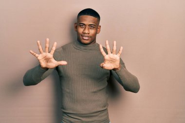Balıkçı yaka kazak giyen genç siyahi bir adam dokuz numaralı parmaklarını gösterip gösterirken kendinden emin ve mutlu bir şekilde gülümsüyor.. 