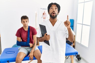 Kırık kolu olan bir adamla birlikte acı giderme kliniğinde çalışan genç İspanyol adam şaşkın şaşkın yukarı bakıyor ve parmaklarıyla işaret ediyor ve kollarını kaldırıyor.. 