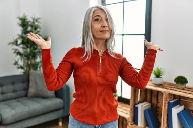 Orta yaşlı, gri saçlı, sıradan kıyafetler giyen bir kadın. Hiçbir şeyden haberi olmayan, elleri ve kolları havada, kafası karışmış bir kadın. şüphe kavramı. 