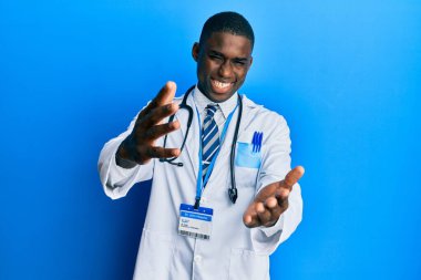 Genç Afrikalı Amerikalı doktor üniforması giymiş kameraya sarılıp gülümsüyor. Mutluluğu kucaklayan neşeli bir ifade.. 