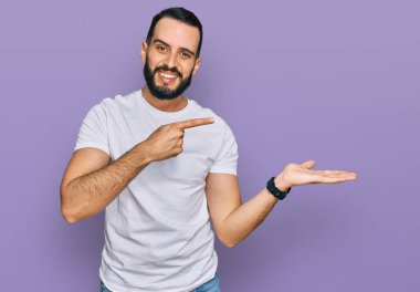 Sakallı, sıradan beyaz tişörtlü genç adam kameraya hayret ediyor ve gülümsüyor.. 