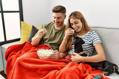 Genç İspanyol çift gülümsüyor. Mutlu bir şekilde kanepede oturmuş, evde köpekleriyle film izliyorlar..