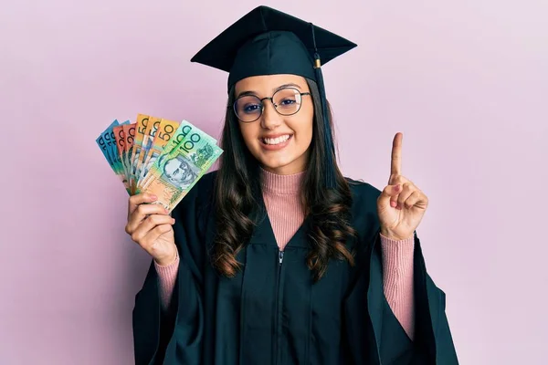 Young Hispanic Woman Wearing Graduation Uniform Holding Australian Dollars Smiling — Stock Photo, Image