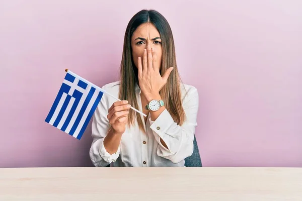 Mujer Hispana Joven Sosteniendo Bandera Griega Sentada Mesa Cubriendo Boca —  Fotos de Stock