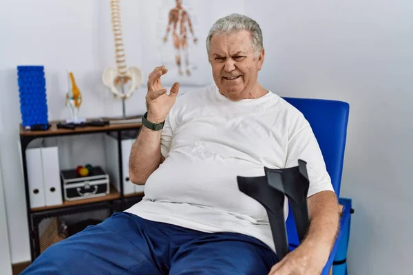 Homem Caucasiano Sênior Clínica Fisioterapia Segurando Muletas Loucas Loucas Gritando — Fotografia de Stock