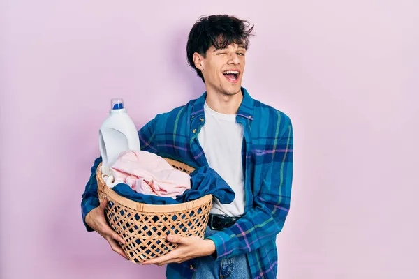 Handsome hipster young man holding laundry basket and detergent bottle winking looking at the camera with sexy expression, cheerful and happy face. — Stock Image Handsome Hipster Young Man Holding Laundry Basket Detergent Bottle Winking Stock Image