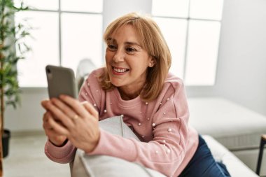 Orta yaşlı sarışın kadın akıllı telefon kullanıyor. Evdeki kanepede oturuyor..