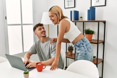 Genç beyaz çift dizüstü bilgisayar kullanıyor ve kahve içiyor. Evdeki masada oturuyorlar..