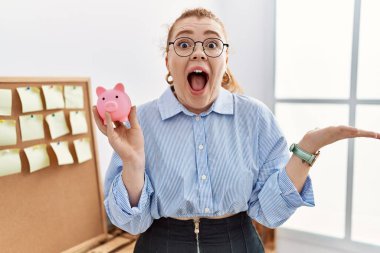 Ofiste domuz kumbarasını tutan genç kızıl saçlı kadın zaferi mutlu bir gülümseme ve ellerini kaldırarak kazanan yüz ifadesiyle kutluyor. 