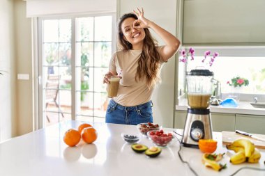 Güzel, genç, esmer bir kadın mutfakta bir bardak smoothie içerken mutlu bir şekilde gülümsüyor. 