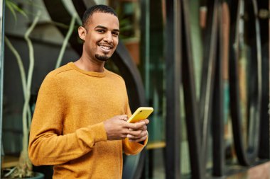 Genç Afrikalı Amerikalı adam şehirde akıllı telefon kullanarak mutlu bir şekilde gülümsüyor..