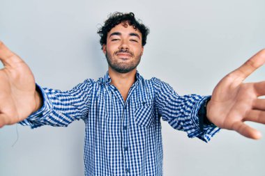 Gündelik giysiler giymiş genç İspanyol adam kameraya bakıp sarılmak için kollarını açmış gülümsüyor. Mutluluğu kucaklayan neşeli bir ifade.. 