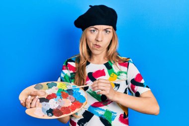 Kafkasyalı genç kadın sanatçı elinde boya fırçası ve palet tutarken şüpheci ve gergin, sorun yüzünden somurtuyor. negatif kişi. 