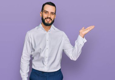 Sakallı, iş gömleği giyen, neşeli gülümseyen, eli avucunda kameraya bakan genç bir adam.. 