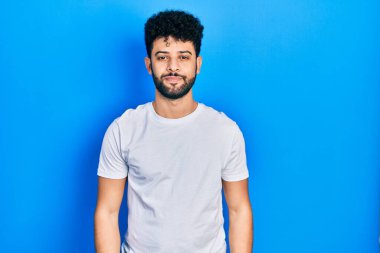 Sakallı, sıradan, beyaz tişörtlü, yüzünde ciddi bir ifadeyle rahatlamış genç bir Arap. Basit ve doğal bir şekilde kameraya bakıyorsun.. 