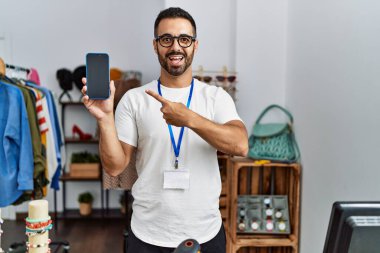 Satış mağazasında müdür olarak çalışan sakallı genç İspanyol adam elinde akıllı bir telefonla mutlu bir şekilde elini ve parmağını göstererek gülümsüyor. 