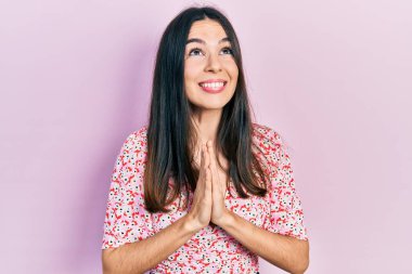 Genç esmer kadın, yaz elbisesi giyip elleri bağlı yalvarıyor ve dua ediyor. Yüzünde umut ifadesi var. Çok duygusal ve endişeli. Yalvarıyor. 