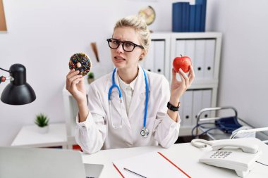 Klinikte elinde kırmızı elmayla donut tutan genç bir doktor. Hiçbir şeyden habersiz ve kafası karışık. şüphe kavramı. 