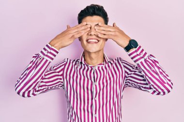 Latin asıllı genç adam günlük kıyafetler giyiyor. Gözleri kapatıyordu. Neşeli ve komik gülümsüyordu. kör kavram. 