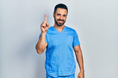Sakallı, mavi erkek hemşire üniforması giyen yakışıklı İspanyol bir numarayı gösterip işaret ederken kendinden emin ve mutlu gülümsüyor.. 