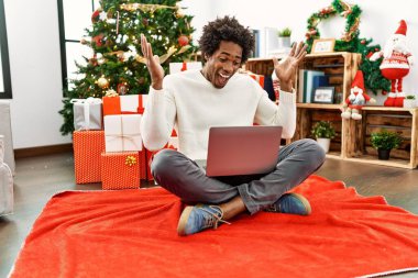 Genç Afro-Amerikalı adam dizüstü bilgisayar kullanıyor. Noel ağacının yanında oturmuş başarının çılgınlığını kutluyor. Kolları havada ve gözleri kapalı heyecanlı bir şekilde bağırıyor. Kazanan konsept 