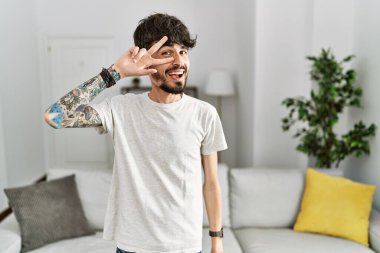 Evdeki oturma odasında sakallı İspanyol bir adam barış sembolü yapıyor. Yüzünde parmakları, gülümseyerek zaferini gösteriyor. 