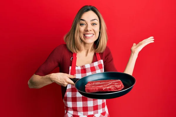 Νεαρή Καυκάσια Ξανθιά Γυναίκα Φορώντας Ποδιά Μαγειρέματος Κρέας Στο Τηγάνι — Φωτογραφία Αρχείου