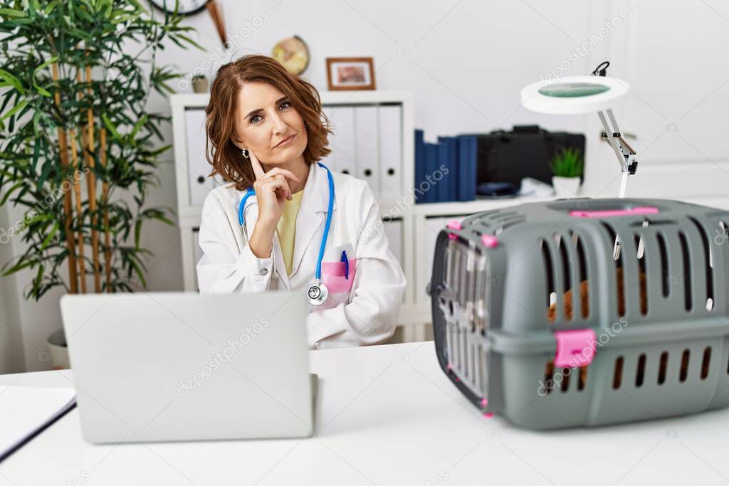 Middle age veterinarian woman working at pet clinic with hand on chin thinking about question, pensive expression. smiling with thoughtful face. doubt concept. 