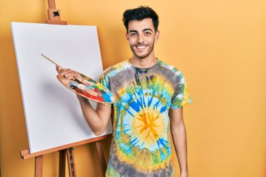 Genç, İspanyol asıllı bir adam yüzünde mutlu ve havalı bir gülümsemeyle paletle resim çiziyor. şanslı kişi. 