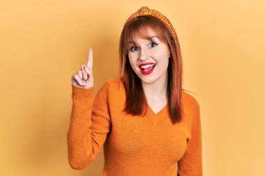 Kızıl saçlı, turuncu süveter giyen genç bir kadın kendini beğenmiş ve mutlu bir şekilde gülümserken bir numarayı işaret ediyor.. 
