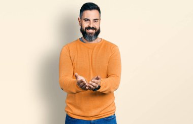 Kış süveteri giyen, elleri avuçlarında gülümseyen ve el hareketi yapan İspanyol sakallı bir adam. Bekleme ve koruma 