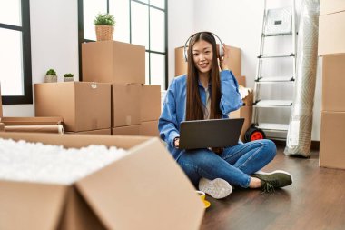 Dizüstü bilgisayar ve kulaklık kullanan Çinli genç bir kız yeni evinde yerde oturuyor.